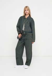 Denim jakna in ujemajoče široke hlače v bledi temno zeleni odtenku. Jakna ima zaklep na gumb in žepek, gladka tekstura.