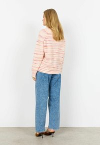 Persoană cu păr blond poartă un pulover roz cu dungi, jeansi largi albaștri și pantofi maro cu toc înalt, stând într-un perete alb.