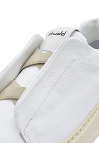 Sneaker bianca in pelle con una fascia elastica beige sopra la linguetta. Presenta una superficie testurizzata e un logo sottile sul tallone. Suola color crema.