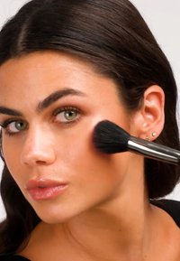 Makeupkost med myke, svarte børster påfører blush på en lys hudtone. Leppene er glansfulle, og håret er glatt og mørkt. Øredetaljer er synlige.