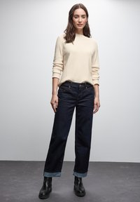 Jeans in denim scuro con gamba affusolata e risvolti, abbinati a un top a maniche lunghe color beige chiaro. Stivali neri alla caviglia completano il look.