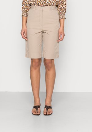 Femme portant un short beige mi-long, un haut à manches longues à motifs et des sandales noires à bouts ouverts, debout devant un fond uni.