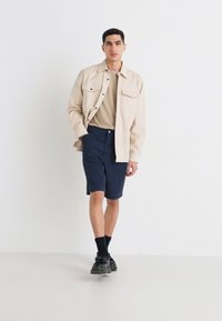 Beige button-up shirt over a tan t-shirt, paired with navy shorts. Worn with black socks and chunky shoes. Plain background.