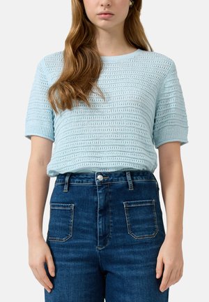 Vrouw draagt lichtblauw gebreid topje met korte mouwen, ingestopt in hooggetailleerde donkerblauwe spijkerbroek met voorzakken.