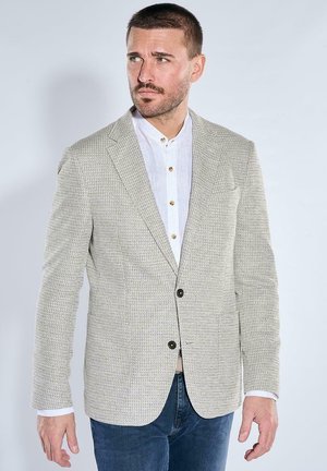 Man with short hair and beard wearing a textured beige blazer over a white buttoned shirt and dark blue jeans, looking to the side.