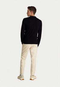Junger Mann steht, trägt ein schwarzes Langarmshirt, beige Hose, weiße Socken und beige Turnschuhe mit braunen Sohlen, und steht seitlich vor einem weißen Hintergrund.
