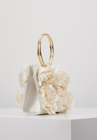 Sac à main beige avec accents floraux en 3D, fabriqué en tissu. Il présente un design structuré et deux poignées circulaires en or.