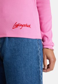 Nahaufnahme einer Person in einem pinkfarbenen Ripp-Langarmshirt mit roter „Lieblingstück“-Stickerei und blauen Jeans, die Hand entspannt an der Seite.