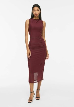 Femme aux longs cheveux noirs portant une robe midi bordeaux sans manches et des sandales noires à talons hauts, debout devant un fond blanc.
