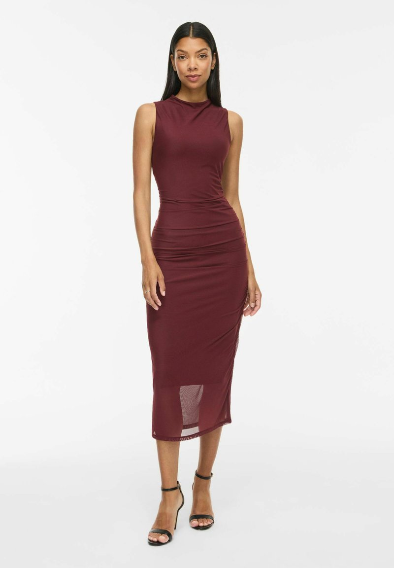 Femme aux longs cheveux noirs portant une robe midi bordeaux sans manches et des sandales noires à talons hauts, debout devant un fond blanc.