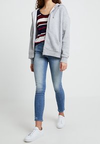 Šedá mikina na zip s přední kapsou, nošena přes červené a bílé pruhované tričko. V kombinaci s přiléhavými modrými džíny a bílými teniskami.