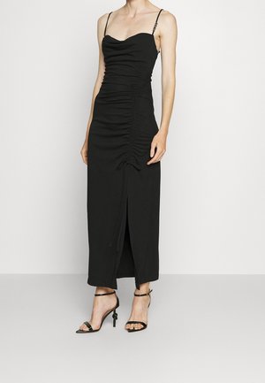 Femme portant une robe midi noire ajustée avec des détails froncés, fines bretelles, une fente avant et des sandales noires à talons avec brides à la cheville.