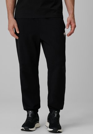 Mężczyzna ubranego w czarne spodnie dresowe i czarne sportowe buty, stojący na jednolitym tle z rękami swobodnie opuszczonymi wzdłuż ciała.