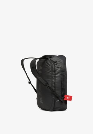 Person iført en beige frakke og sorte bukser, bærer en stor sort Mammut rygsæk set bagfra mod en ensfarvet grå baggrund.