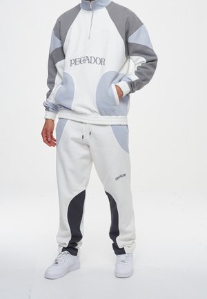 Hombre vistiendo un chándal bloqueado en colores blanco, gris y negro con el texto "PEGADOR" en la sudadera y los pantalones, de pie con una mano en el bolsillo.