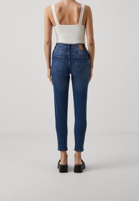 Vysoké džíny z tmavomodrého denimového materiálu se slim fit střihem. Mají dvě zadní kapsy a malou hnědou koženou nášivku na pase.