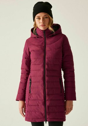 Kobieta w długim, bordowym pikowanym płaszczu z kapturem i czarnej czapce, stojąca na tle jednolitego jasnego tła.