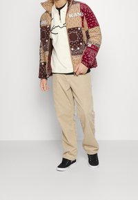 Lappat jacka i nyanser av brunt och rött med bandana-mönster, tillsammans med en krämfärgad skjorta och beige kordflöjlsbyxor. Svarta sneakers.