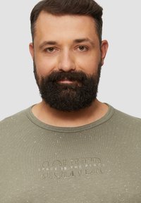 Olivgrünes T-Shirt mit Rundhalsausschnitt, das einen strukturierten Druck mit der Aufschrift "SOLVER SPACE IS THE PLACE" und dezenten weißen Sprenkeln aufweist.