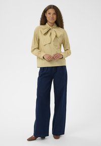 Femme avec des cheveux bouclés portant un chemisier rayé jaune avec un grand nœud et un jean bleu foncé à jambes larges, se tenant contre un fond blanc.