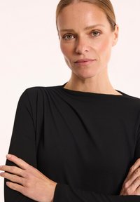 Czarny top z długim rękawem i okrągłym dekoltem, gładka tekstura, dopasowany krój i delikatny połysk, noszony z krzyżującymi się rękami na neutralnym tle.