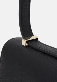 Černá kožená kabelka zblízka ukazující texturovaný povrch, elegantní design a zlatý kovový dekorativní prvek spojující rukojeť.