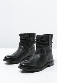 Botas de cuero negro hasta el tobillo con un diseño desarenado, punta redondeada, tacón bajo y detalles de costura sutiles. Textura suave con un hardware mínimo.