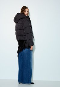 Veste noire matelassée avec capuche, présentant un design en patchwork. Assortie à un jean en denim bleu à jambes larges et un haut noir orné de dentelle.