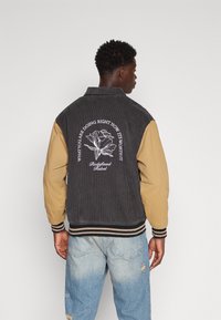Veste en velours côtelé et tissu tissé avec un corps noir et des manches beige, présentant une broderie florale au dos et des poignets côtelés rayés.