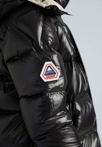 Manche d'une veste matelassée noire brillante avec un écusson hexagonal "JACKIT" en rouge, blanc et bleu sur le haut du bras, sur un fond clair.