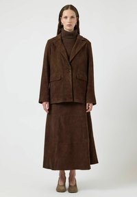 Blazer in suede marrone con un singolo bottone, abbinato a una lunga gonna coordinata. Tessuto testurizzato, design semplice e taglio al ginocchio. Calzature neutre.