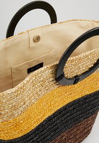 Woven straw tote bag featuring horizontal stripes in beige, yellow, black, and brown. Black circular handles and beige fabric lining inside.