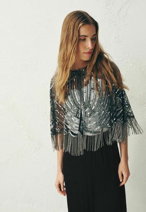 Frau mit langen Haaren, die einen schwarzen, mit Pailletten besetzten Umhang mit Fransen über einem grauen Oberteil und einem schwarzen Rock trägt, schaut nach links unten.