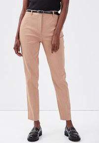 Ceinture noire sur un pantalon beige taille haute porté avec des mocassins noirs à clous, haut noir sans manches court, mannequin se tenant contre un fond blanc.