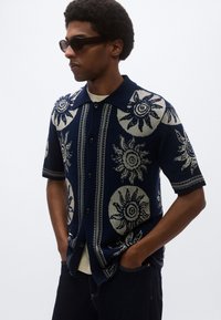 Λεπτή πλεκτή μπλούζα ναυτικού μπλε με κοντά μανίκια, διακοσμημένη με κρεμ μοτίβα ήλιου, κλείνει με κουμπιά μπροστά, έχει επίπεδη γιακά και αντιθετικές υφασμάτινες ρίγες.