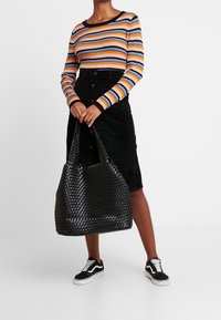 Sac fourre-tout tissé noir avec surface texturée, anses larges et design spacieux, assorti à un haut côtelé rayé multicolore et une jupe noire.