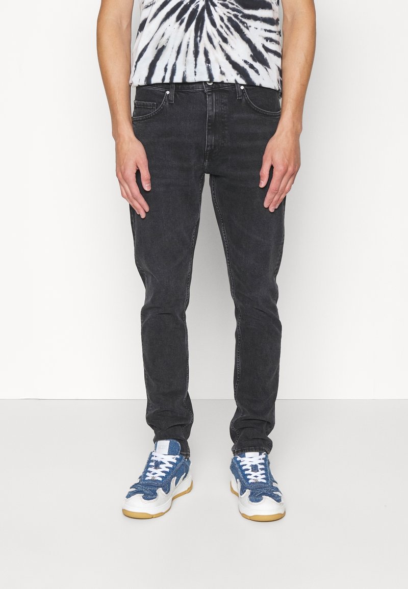 Mužský model stojí v tričku s batikovaným vzorem, černých džínách a modrých teniskách. Pozadí je čistě bílé, což ještě více zdůrazňuje jeho oblečení.