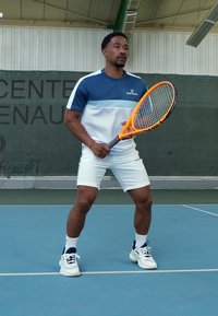 Tennisser in een blauw-witte sportshirt en witte shorts, met een oranje tennisracket in de hand, staand op een blauwe tennisbaan.