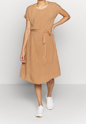 Vrouw draagt een bruine jurk tot op de knie met witte verticale strepen, een gestrikte tailleband en witte sneakers, staand met één hand op haar heup.