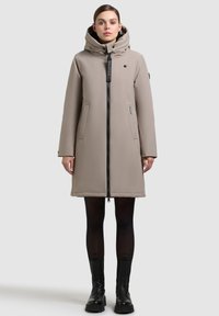 Dlhý béžový puffrový kabát s vysokým golierom a odnímateľnou kapucňou. Obsahuje predný zips, dva bočné vrecká a logo nášivku na hrudi.