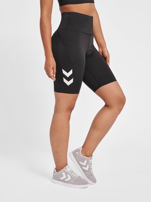 Donna che indossa pantaloncini da bici neri a vita alta con loghi bianchi a chevron e scarpe da ginnastica grigie con design a chevron coordinato, in piedi su sfondo bianco.