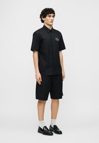 Νεαρός άνδρας που φοράει μαύρο κοντομάνικο πουκάμισο με κουμπιά και σορτς μέχρι το γόνατο, με άσπρες κάλτσες και μαύρα δερμάτινα loafers, όρθιος πλάγια.