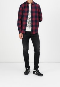 Camisa de cuadros roja y azul marino abotonada sobre una camiseta gráfica blanca, combinada con jeans negros y zapatillas negras con rayas blancas.