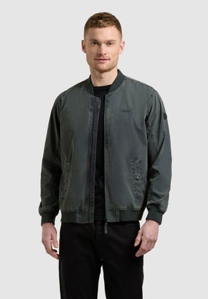 Muž v tmavozelenej bombera bunde cez čierne tričko, stojaci pred jednoduchým svetlosivým pozadím.