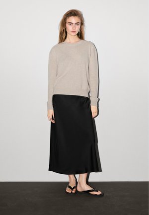 Maglione in maglia beige, gonna maxi in satin nera e infradito nere. La modella è in piedi contro uno sfondo neutro. Design semplice con texture morbide.