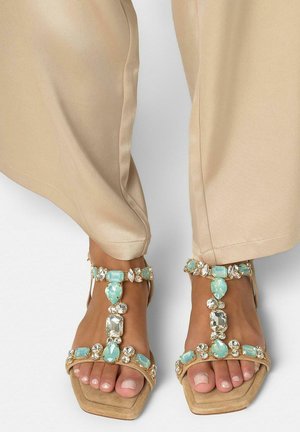 Pieds portant un pantalon beige et des sandales à bouts ouverts décorées de grandes pierres précieuses bleu clair et transparentes sur un fond clair.