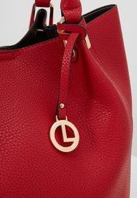 Sac à main en cuir texturé rouge avec bordure noire et pendentif logo circulaire doré suspendu à la sangle de la poignée.