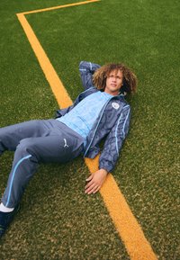 Completo sportivo Puma in blu chiaro e grigio scuro; giacca con motivo grafico e accenti blu. Adagiato su un prato verde accanto a una linea gialla.