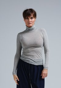 Sivý pulóver s dlhým rukávom a stojačikom, s priliehavým strihom, vyrobený z jemného materiálu, má drapovaný lem a príjemnú textúru. V páre s tmavomodrými nohavicami.