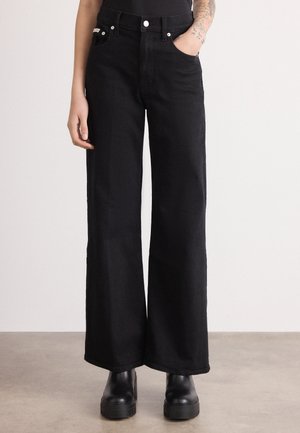 Personne portant un jean noir taille haute à jambes larges et des bottes plateformes noires, avec une main dans la poche et un avant-bras tatoué visible.